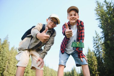 Zengin bir ormanda yürüyüş yaparken, sırt çantalı ve dürbünlü iki neşeli çocuk yaz tatilinin tadını çıkarıyor ve pozitifliklerini dile getiriyorlar.