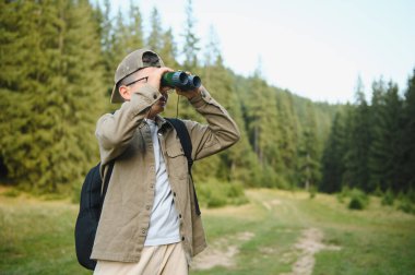 Orman yolunda yürürken dürbün kullanan meraklı çocuk doğa macerası sırasında doğanın güzelliğinin tadını çıkarıyor.