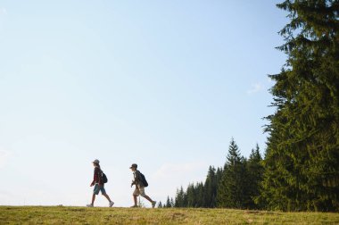 İki genç yürüyüşçü güneşli bir günde dağ manzarasında yürüyüş yapıyor, sırt çantaları taşıyor ve günlük giysiler giyiyorlar.