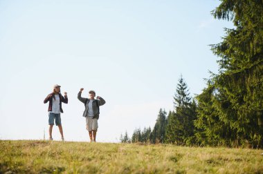 İki genç yürüyüşçü, yemyeşil bir ormanla çevrili bir dağın zirvesine ulaştıktan sonra kutlama için kollarını kaldırıyorlar.