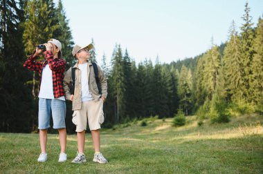 Kaşif kıyafetleri giyen iki çocuk ormanda yürüyüş yaparken, biri dürbünle etrafı gözetliyor.
