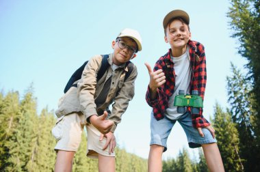 Dürbünlü ve sırt çantalı iki neşeli çocuk yemyeşil bir ormanda yürüyüş yaparken başparmaklarını gösteriyorlar.