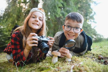İki genç turist ormanda çimlerin üzerinde uzanmış büyüteç ve kamerayla mantarları izliyorlar.