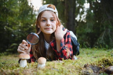 Doğayı keşfeden meraklı kız, yemyeşil bir ormanda yürüyüş yaparken büyüteçle mantarları gözlemliyor.