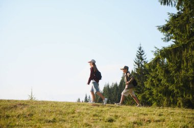İki genç izci, güneşli bir günde, yüksek ağaçlarla çevrili yemyeşil bir çayırda manzaralı bir yürüyüşün tadını çıkarıyorlar.