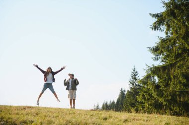 İki genç yürüyüşçü ormanda neşeyle zıplıyor, canlı yeşil ve güneşli gökyüzünün ortasında birlikte maceralarını kutluyorlar.