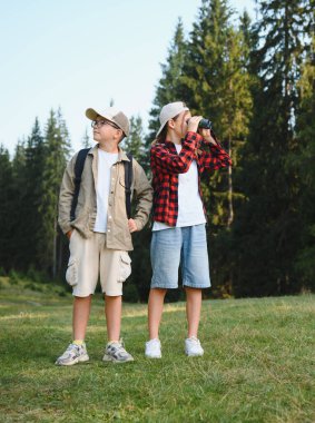 İki genç yürüyüşçü, bir erkek ve bir kız, yemyeşil bir ormanı keşfediyorlar, kız dürbünle etrafı gözetlerken kız da onunla birlikte.