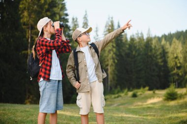 Doğayı keşfeden sırt çantalı iki genç turist, dürbünle bakan kız, uzaklığı gösteren oğlan, orman manzarası ile çevrili.