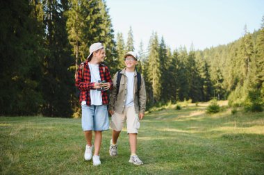 Sırt çantalı iki genç yürüyüşçü, yemyeşil yemyeşil ve sıcak güneş ışığıyla çevrili canlı bir orman çayırında güneşli bir yürüyüş yapıyorlar.