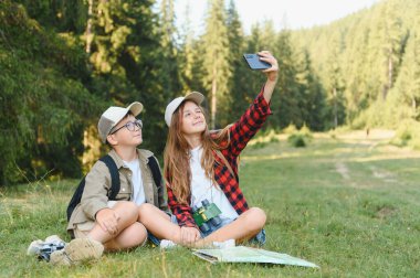 İki genç izci, bir yürüyüş sırasında ormanda çimlerin üzerinde dinlenirken akıllı bir telefonla selfie çekiyorlar.