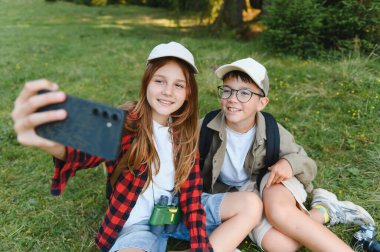 Kaşif kıyafeti giymiş iki çocuk ormanda yürüyüş yaparken akıllı telefonlarıyla selfie çekiyorlar.