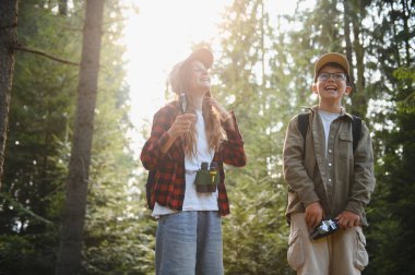 Güneşli bir ormanda yürüyüş yapan iki neşeli çocuk sırt çantaları, dürbünler ve bir kamera taşıyarak ormanı keşfediyorlar.