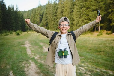 Büyüteci ve dürbünü olan mutlu bir çocuk ormanda gezintiden zevk alıyor, bir keşfi kollarını açıp kutluyor.