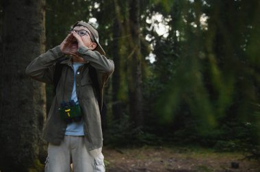 Genç izci gözlüklü ve dürbünlü bir çocuk sık bir ormanda yüksek sesle bağırıyor, muhtemelen kaybolmuş ve yardım istiyor.