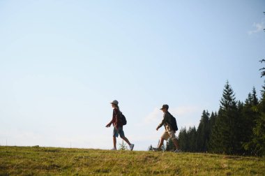 Sırt çantalarıyla çimenli bir tepede yürüyen iki genç yürüyüşçü güneşli bir günün tadını çıkarıyorlar. Manzaralı bir doğa yolunu keşfediyorlar.