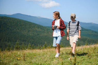 İki genç turist çimenli bir dağ yamacında yürüyor, canlı yaz manzarasında ıslanıyorlar ve açık havanın tadını çıkarıyorlar.
