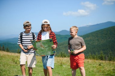 Çocuklar yaz tatillerinde güzel bir dağ manzarasında yürüyüş yaparken haritaya bakıp dürbün kullanıyorlar.