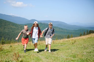 Üç neşeli çocuk el ele tutuşup çimenli bir dağ yamacında koşuyor ve güzel açık havada yaz tatilinin tadını çıkarıyorlar.