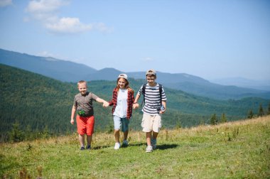 Güneşli bir yaz günü, neşeli bir şekilde el ele tutuşan üç çocuk güneşli dağlarda yürüyüş yapıyor, macera ve doğa ile kucaklaşıyorlar.