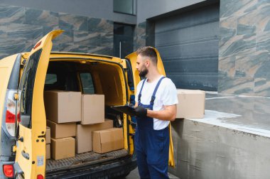 Tulum ve eldiven giyen kurye, sarı bir minibüste karton kutuları kontrol ediyor, elinde kağıt ve kalem tutuyor.