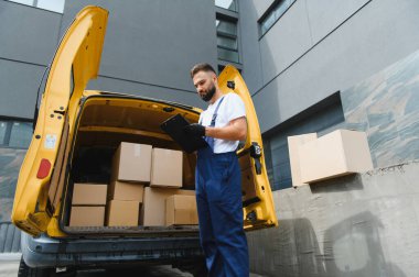 Eldiven takan ve karton kutuları işaretleyen kurye, sarı bir minibüste duruyor, doğru teslimatı sağlamak için bir dosya kullanıyor.