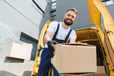 Eldiven takan teslimatçı, elinde bir karton kutu tutarken gülümseyen ve parlak sarı bir minibüsten paketleri boşaltan adam.
