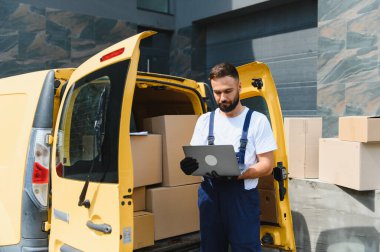 Dağıtılmaya hazır paketlerle dolu sarı bir minibüsün yanına park edilmiş bir dizüstü bilgisayardaki siparişleri kontrol eden kurye.