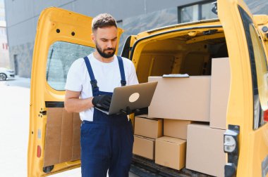 Kurye, karton kutularla dolu minibüsün yanındaki dizüstü bilgisayarda siparişleri kontrol ediyor. Şehir sokaklarında paket teslimatını yönetiyor.