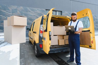 Kargo işçisi, karton kutularla dolu sarı minibüsünün yanındaki dizüstü bilgisayarda siparişleri kontrol ediyor.