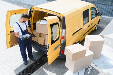 Kurye, sokağın içinde ve dışında karton kutular olan sarı teslimat minibüsünü açmak için dizüstü bilgisayar kullanıyor.