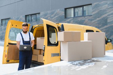 Kurye, karton kutularla dolu sarı minibüsünün yanındaki dizüstü bilgisayardan siparişleri kontrol ediyor.