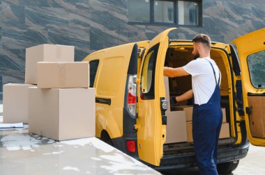 Tulum giyen kurye, modern bir binanın yakınına park edilmiş sarı bir minibüse karton kutular yüklüyor.