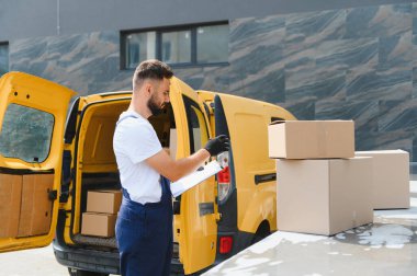 Teslimatçı, şehir merkezinde sarı bir minibüsün yanında dururken karton kutuları işaretliyor.