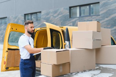 Teslimatçı, karton kutuları kontrol ediyor. Sarı bir minibüsün üstünde, nakliye ve lojistik işleriyle uğraşıyor.