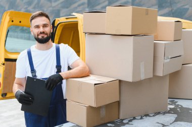 Karton kutular ve parlak sarı bir teslimat minibüsünün yanında konumlandırılmış paketleri denetleyen kurye