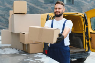 Eldivenli kurye, müşteriye karton bir kutu veriyor. Arkasında yığılı paketler ve sarı bir minibüs var.