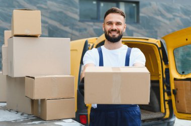 Bir paket taşıyan kurye, karton kutularla dolu sarı bir teslimat aracının yanında gülümseyerek nakledilmeye hazır.