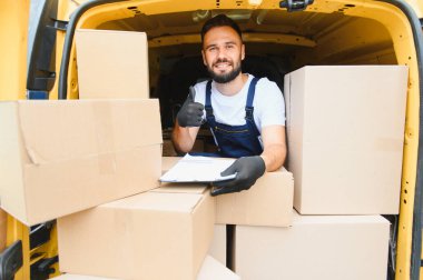 Bir kargo aracının içinde karton kutularla çevrili bir pano tutarken kurye başparmaklarını kaldırıyor