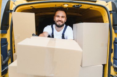 Bir karton kutuyu paketlerle dolu bir kamyonetin içinde tutarken kurye gülümsüyor. Etkili lojistik ve servis ruhunu somutlaştırıyor.