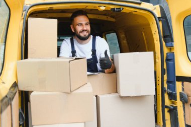 Karton kutularla dolu sarı minibüsünde paketleri teslim etmeye hazırlanırken kurye başparmağını kaldırıyor.