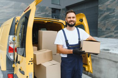 Eldivenli kurye, karton kutularla dolu sarı minibüsünün yanında bir paket tutuyor, gülümsüyor.