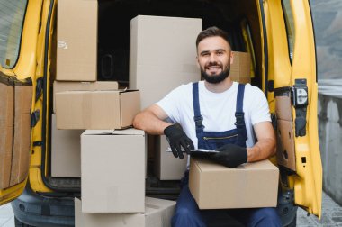 Teslimatçı, karton kutularla dolu bir minibüste kameraya gülümseyen paketlerin listesini kontrol ediyor.