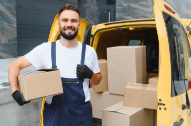 Kurye, sarı teslimat minibüsünün yanında, gülümsüyor ve eldiven ve üniforma giyiyor.