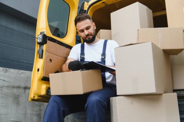 Eldiven giyen ve üniforma giyen bir teslimatçı sarı bir minibüsün arkasında oturup karton kutuları kağıt ve kalemle kontrol ediyor.