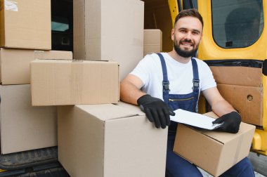 Karton kutularla dolu bir minibüste otururken teslimatçı paketleri kontrol ediyor, gülümsüyor.