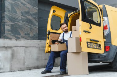 Paket listesini kontrol eden teslimatçı, sarı minibüsünde açık kapılı, karton kutuları teslim etmeye hazır bekliyor.