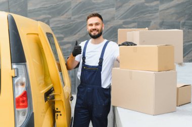 Gülümseyen teslimatçı, karton kutular ve sarı teslimat minibüsünün yanında baş parmağını kaldırıyor.
