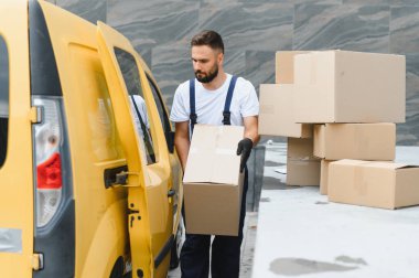 Sarı bir minibüsten paketleri boşaltan kurye, eldiven giyerken karton bir kutu taşıyor.