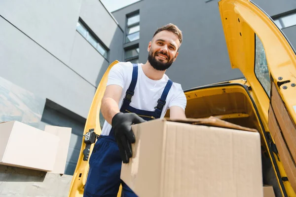 Tulum giymiş, siyah eldivenli gülümseyen teslimatçı, sarı minibüsünün yanında bir karton kutu tutuyor.