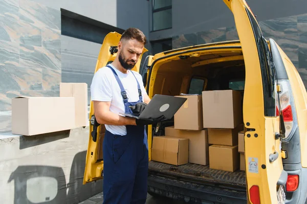 Dağıtım elemanı, karton kutularla dolu sarı minibüsün yanında dizüstü bilgisayar kullanıyor. Nakliye ve lojistik işlerini yönetiyor.